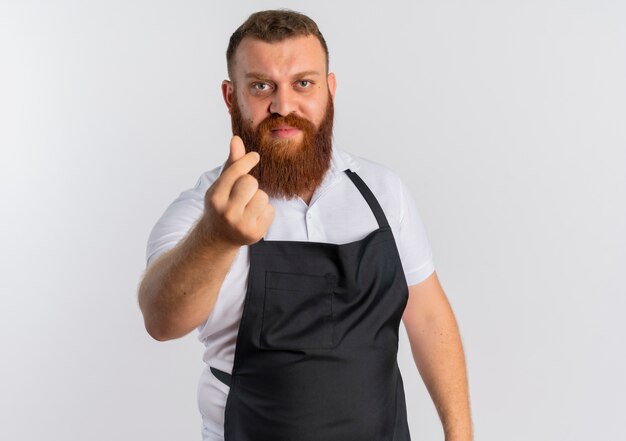 Professional bearded barber man in apron rubbing fingers making money gesture standing over white wall