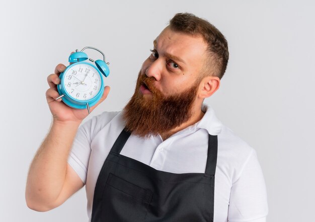 Профессиональный бородатый парикмахер в фартуке показывает будильник со скептическим выражением лица, стоящий над белой стеной