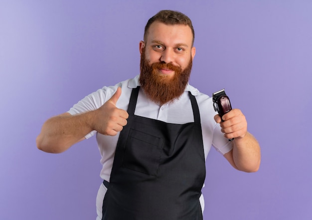 Professional bearded barber man in apron showing hair cutting machine pointing with finger to it smiling showing thumbs up standing over purple wall