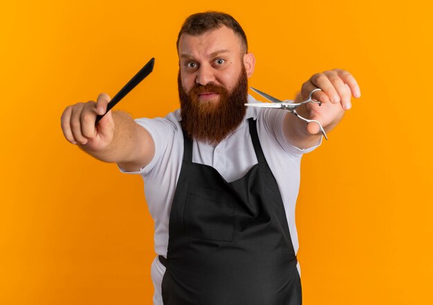 Professional bearded barber man in apron showing scissors and hairbrush looking confused standing over orange wall