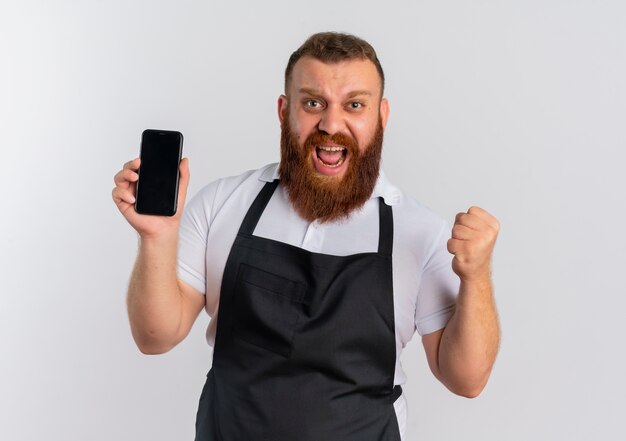 Professional bearded barber man in apron showing smartphone clenching fist happy and excited standing over white wall