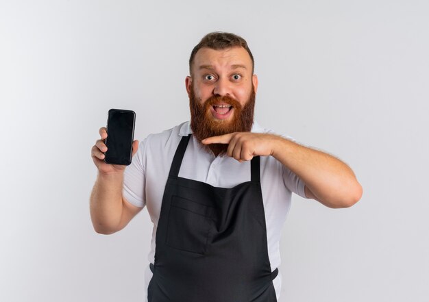 Профессиональный бородатый парикмахер в фартуке показывает смартфон, указывая пальцем на него, улыбаясь счастливым и взволнованным, стоя над белой стеной