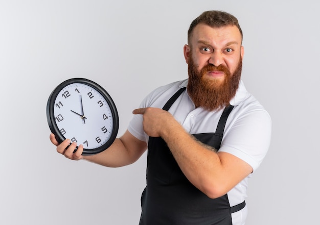 Professional bearded barber man in apron showing wall clock pointing with finger to it displeased standing over white wall