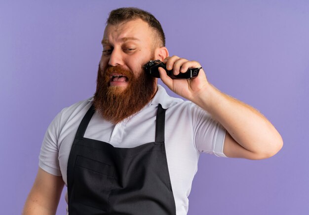 紫色の壁の上に立っている顔の悲しい表情と混同しているように見えるシェービングマシンで彼のひげをトリミングするエプロンのプロのひげを生やした理髪店の男