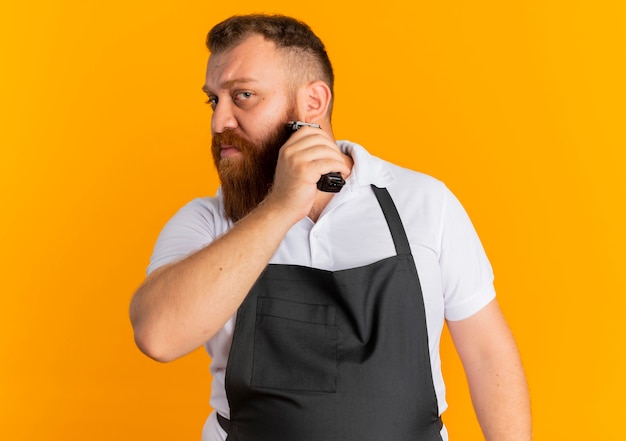 Uomo barbuto professionista del barbiere in grembiule che taglia la barba con la macchina da barba in piedi sopra la parete arancione