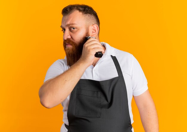 Professional bearded barber man in apron trimming his beard with shaving machine standing over orange wall