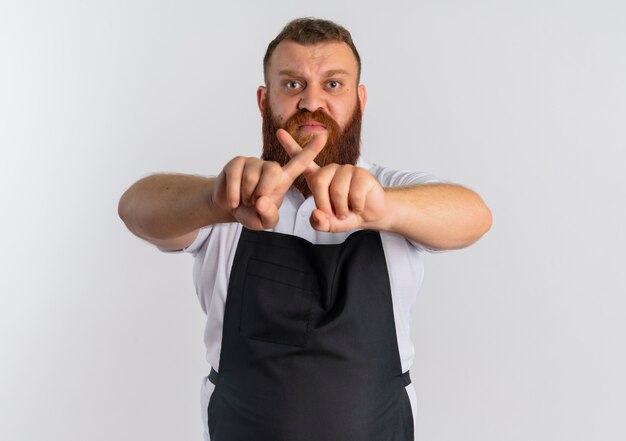Профессиональный бородатый парикмахер в фартуке с сердитым лицом делает знак остановки, пересекающий указательные пальцы, стоящий над белой стеной