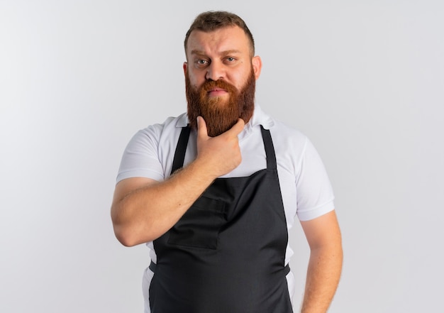Professional bearded barber man in apron with hand on his chin thinking standing over white wall