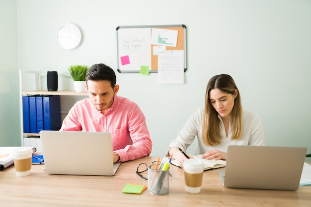 Professional business partners working next to each other at a desk. Caucasian woman writing her task on an agenda and male coworker typing on a laptop