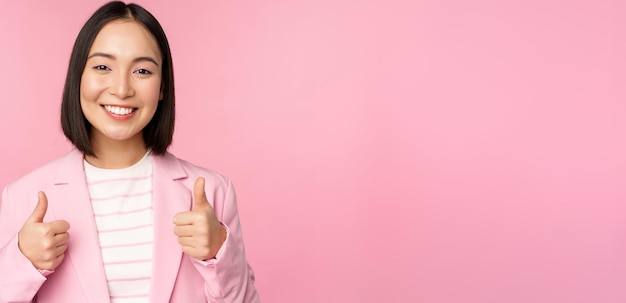 Профессиональная деловая женщина, азиатская корпоративная женщина, показывающая большие пальцы и улыбающаяся похвала и комплимент, стоящая в костюме на розовом фоне