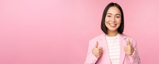 Professional businesswoman asian corporate woman showing thumbs up and smiling praise and compliment standing in suit over pink background