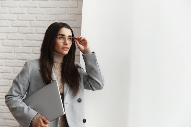 Professional businesswoman in glasses standing near the window with a laptop.