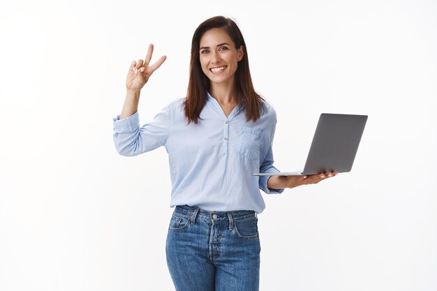 Professional carefree cheerful caucasian 30s woman with tattoo working, feel confident project goes well, show victory peace sign, hold laptop, stand white wall satisfied, assure finish in time
