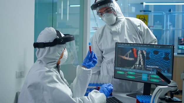 Профессиональные химики в костюме ppe анализируют разработку вакцины, указывая на дисплей компьютера в оборудованной лаборатории