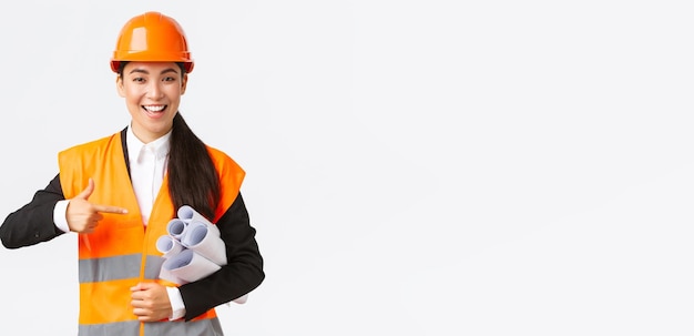 Professional confident asian female architect chief engineer in safety helmet pointing finger at blueprints showing project plan or documents for construction standing white background