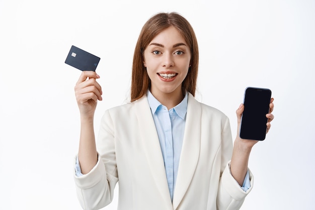 전문 기업 여성, CEO 관리자는 신용 카드와 스마트폰 화면을 보여주며 흰색 벽 위에 비즈니스 정장을 입고 서 있습니다.