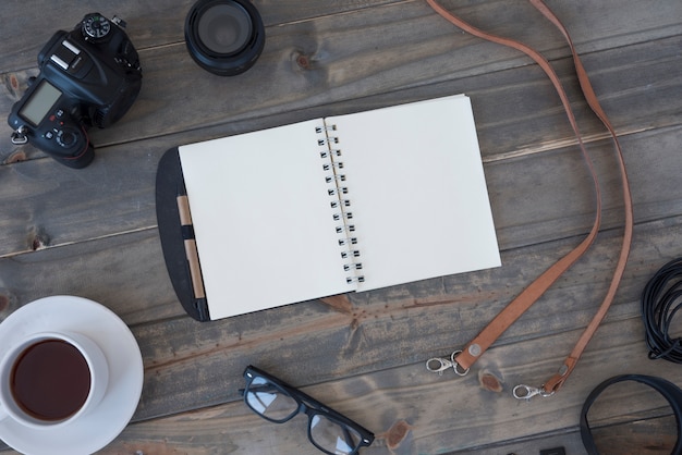 Professional digital camera; cup of coffee; blank spiral notepad; pen; spectacles and cable on wooden table