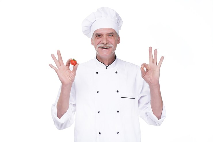 Free Photo professional elderly chef in white uniform and hat, holding tomato while staying on white wall
