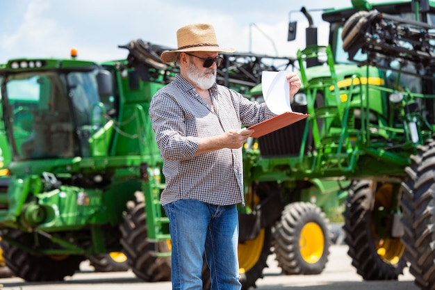 Профессиональный фермер с современным трактором за работой с документами. Смотрит солнышко. Сельское хозяйство, выставки, техника, растениеводство. Старший мужчина возле своей машины.