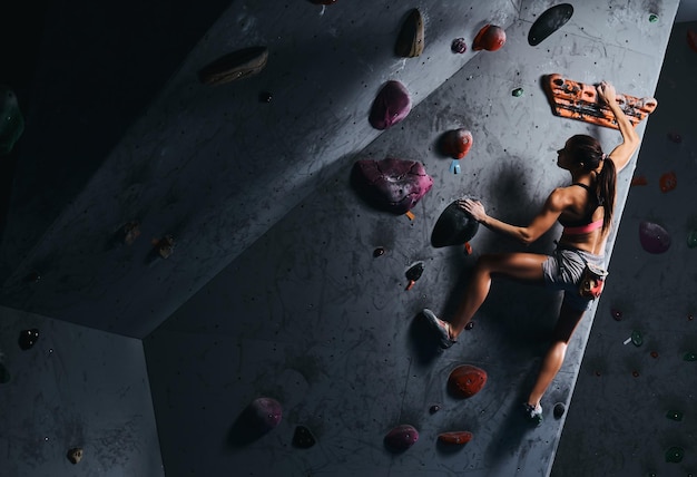 Professional female climber hanging on bouldering wall, practice climbing indoors.