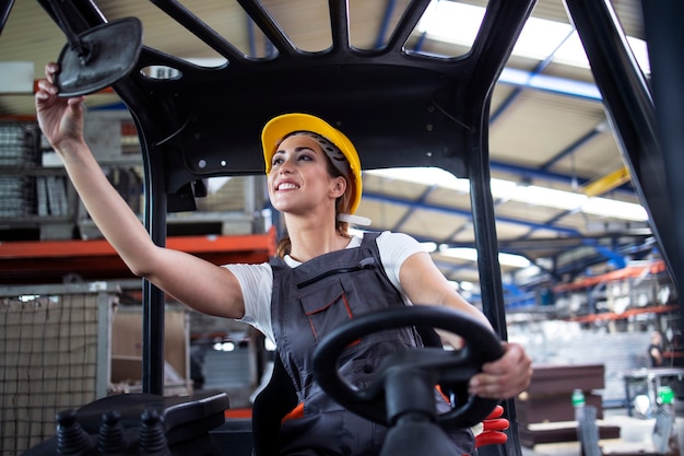 Professional female industrial driver adjusting rear mirrors and operating forklift machine in factory's warehouse