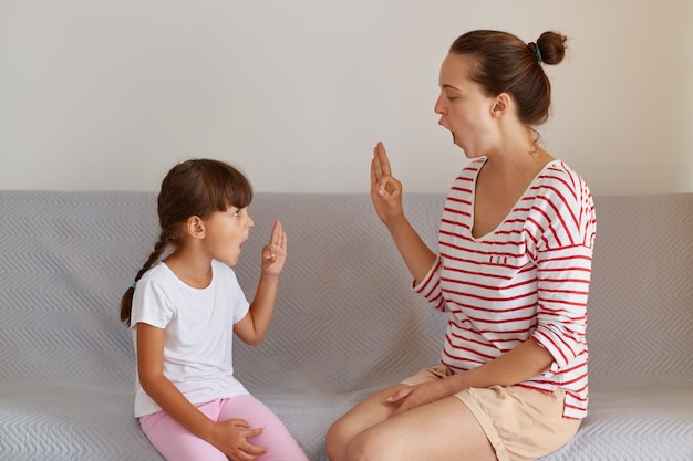 ソファに座っている間、屋内で小さな子供の女の子とのスピーチの欠陥または困難に取り組んでいるプロの女性理学療法士、話すことを改善するための言語レッスンを持っている小さな子供。