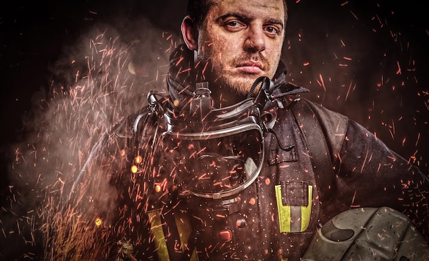 A professional firefighter dressed in uniform holding safety helmet in fire sparks and smoke over a dark background.