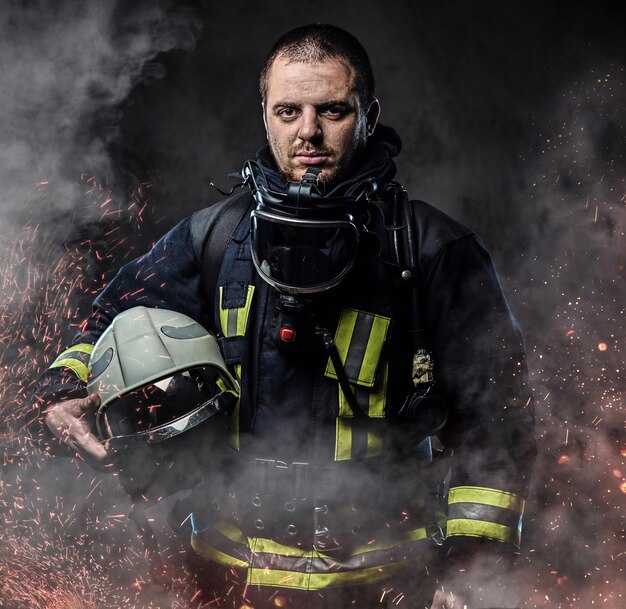 Профессиональный пожарный, одетый в униформу, держит защитный шлем в огненных искрах и дыму на темном фоне.