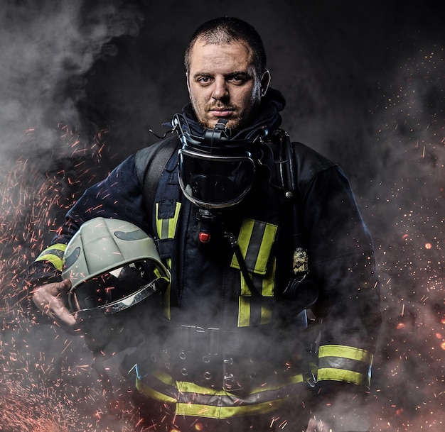 A professional firefighter dressed in uniform holding safety helmet in fire sparks and smoke over a dark background.