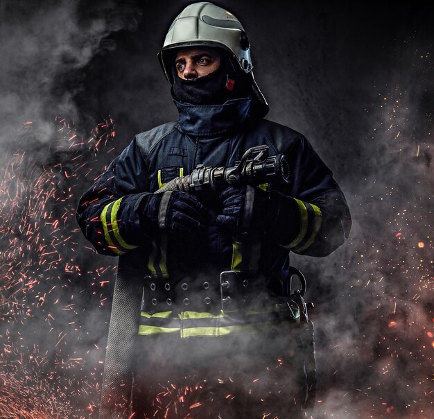 A professional firefighter in uniform holds the fire hose in fire sparks and smoke over a dark background.