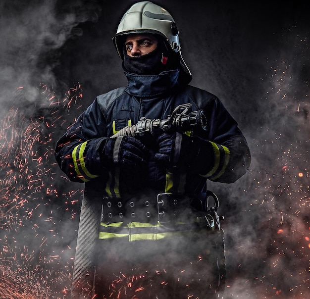 A professional firefighter in uniform holds the fire hose in fire sparks and smoke over a dark background.