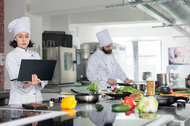 Professional food industry worker with computer searching for gastronomic meal. Female head chef with laptop brainstorming garnish ideas for gourmet dish recipe while standing in restaurant kitchen.