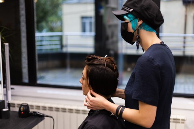 プロの女の子の美容師がクライアントのヘアカットを行います。女の子は美容院でマスクに座っています