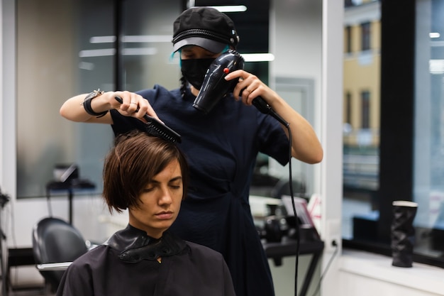 プロの女の子の美容師がクライアントのヘアカットを行います。女の子は美容院でマスクに座っています