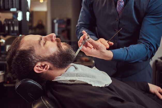 Профессиональный парикмахер моделирует бороду ножницами и расческой в парикмахерской. Фото крупным планом.