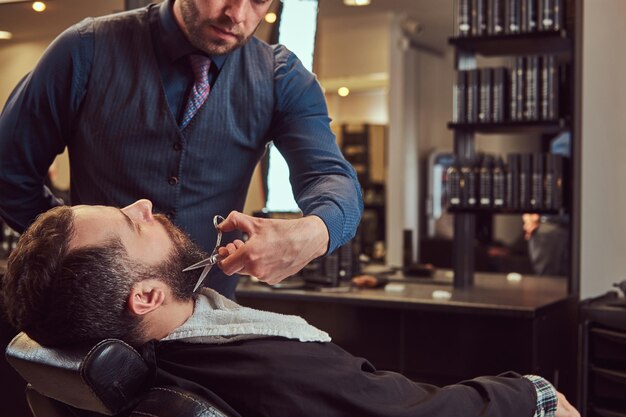 Professional hairdresser modeling beard with scissors and comb at the barbershop. Close-up photo.