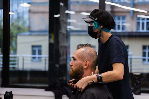 保護フェイスマスクを身に着けているプロの美容師は、美容院でヨーロッパのひげを生やした残忍な男性のために散髪をします