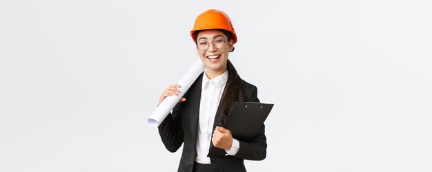 Professional happy asian female architect construction engineer in helmet and business suit holding blueprints and clipboard with building documentation smiling cheerful white background