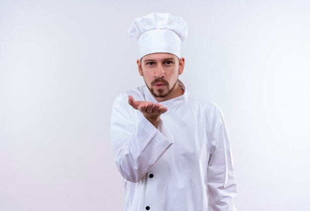 Профессиональный шеф-повар-мужчина в белой униформе и поварской шляпе, дующий воздушный поцелуй с рукой в воздухе, мило стоя на белом фоне