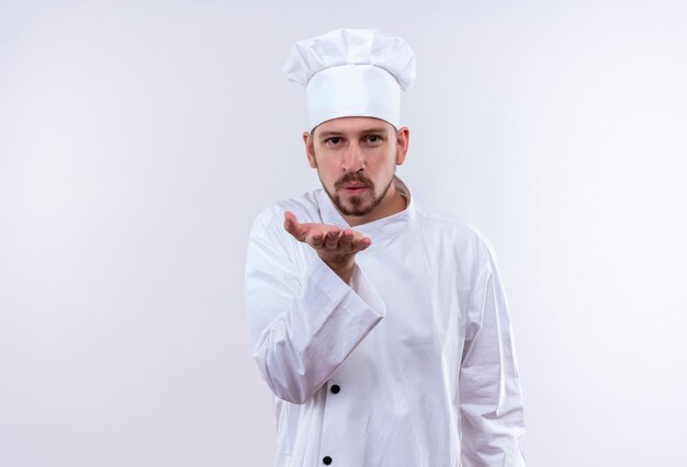 Professional male chef cook in white uniform and cook hat blowing a kiss with hand on air being lovely standing over white background