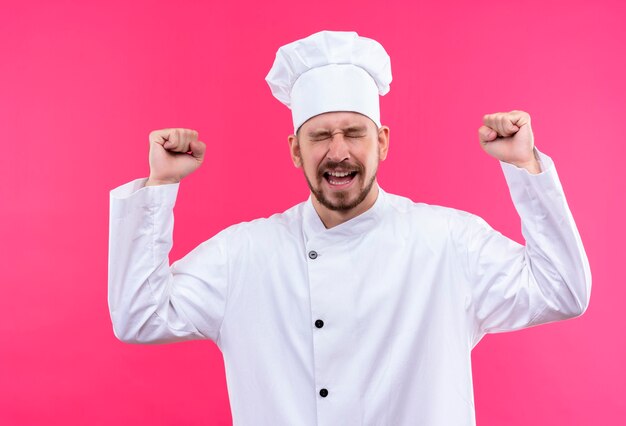 전문 남성 요리사는 흰색 유니폼을 입고 분홍색 배경 위에 서있는 그의 성공을 기뻐하는 닫힌 눈으로 웃고 모자 ceazy 행복 떨림 주먹 요리