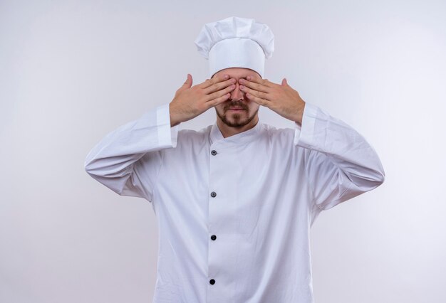 Professional male chef cook in white uniform and cook hat covering eyes with hands standing over white background