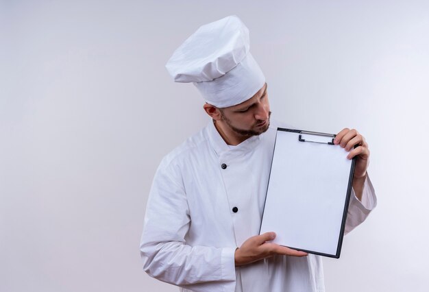 Профессиональный шеф-повар-мужчина в белой форме и поварской шляпе демонстрирует буфер обмена с пустыми страницами, стоящими на белом фоне