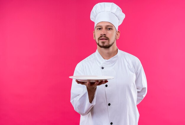 Professional male chef cook in white uniform and cook hat demonstrating a plate looking confident standing over pink background