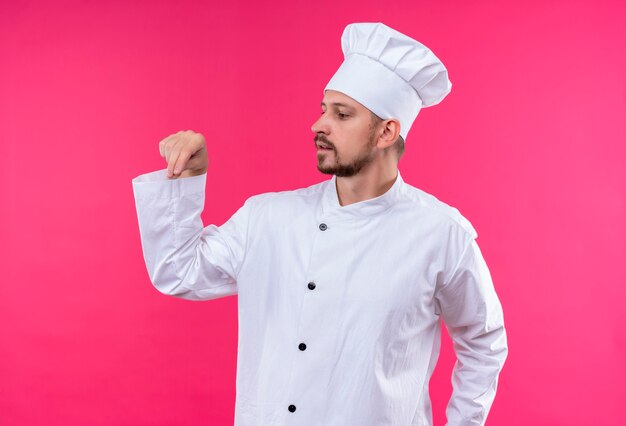Professional male chef cook in white uniform and cook hat gesturing with hand looking confident standing over pink background
