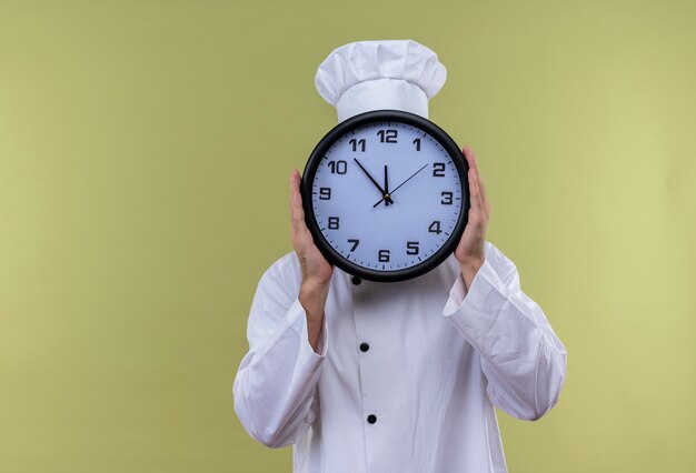 Professional male chef cook in white uniform and cook hat hiding his face behind big clock standing over green background