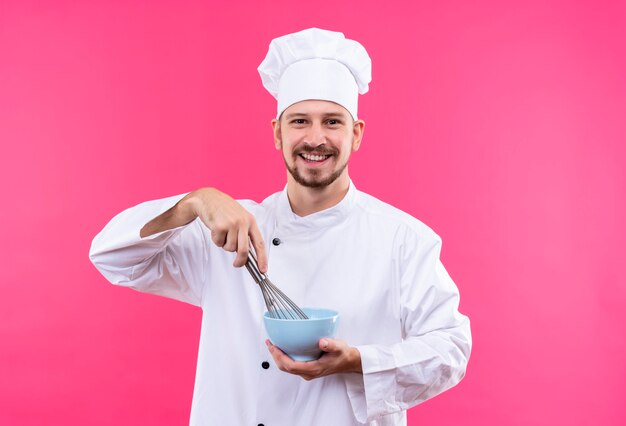 Professional male chef cook in white uniform and cook hat holding a bowl whipping something with whisk smiling cheerfully standing over pink background