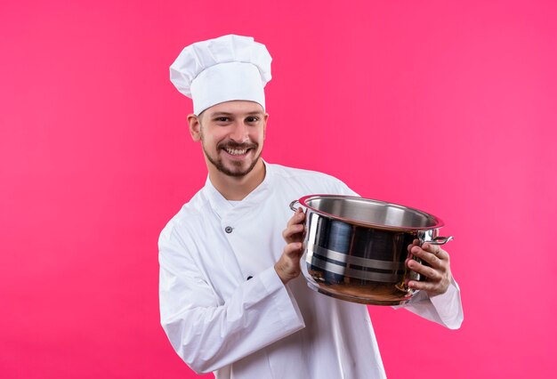 Professional male chef cook in white uniform and cook hat holding an empty pan looking at camera smiling cheerfully standing over pink background