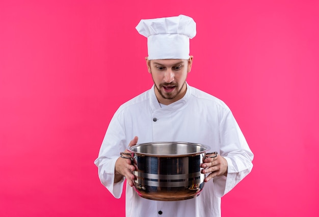 Профессиональный шеф-повар-мужчина в белой форме и поварской шляпе держит пустую кастрюлю, глядя на нее изумленно и удивленно, стоя на розовом фоне