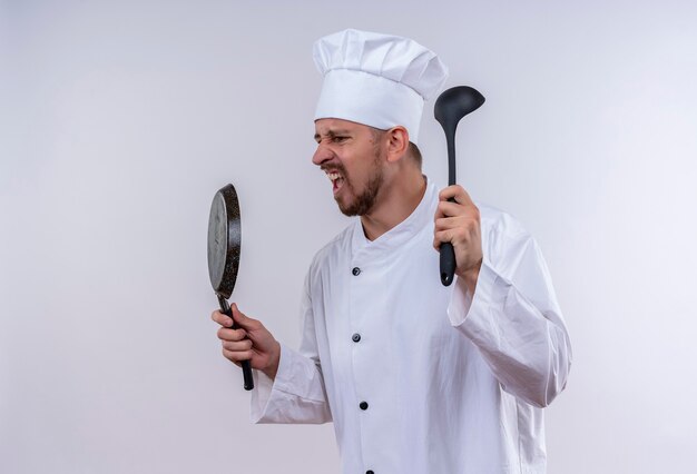 Professional male chef cook in white uniform and cook hat holding frying pan and ladle shouting and yelling with aggressive expression standing over white background